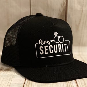 May include: Black trucker hat with a white mesh back. The hat has a white graphic that says "Ring Security" with a diamond ring and two wedding bands.