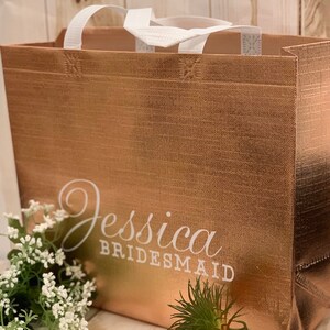 Puede incluir: Una bolsa de tela reutilizable de color oro rosa con asas blancas y el texto "Jessica Bridesmaid" impreso en la parte delantera.