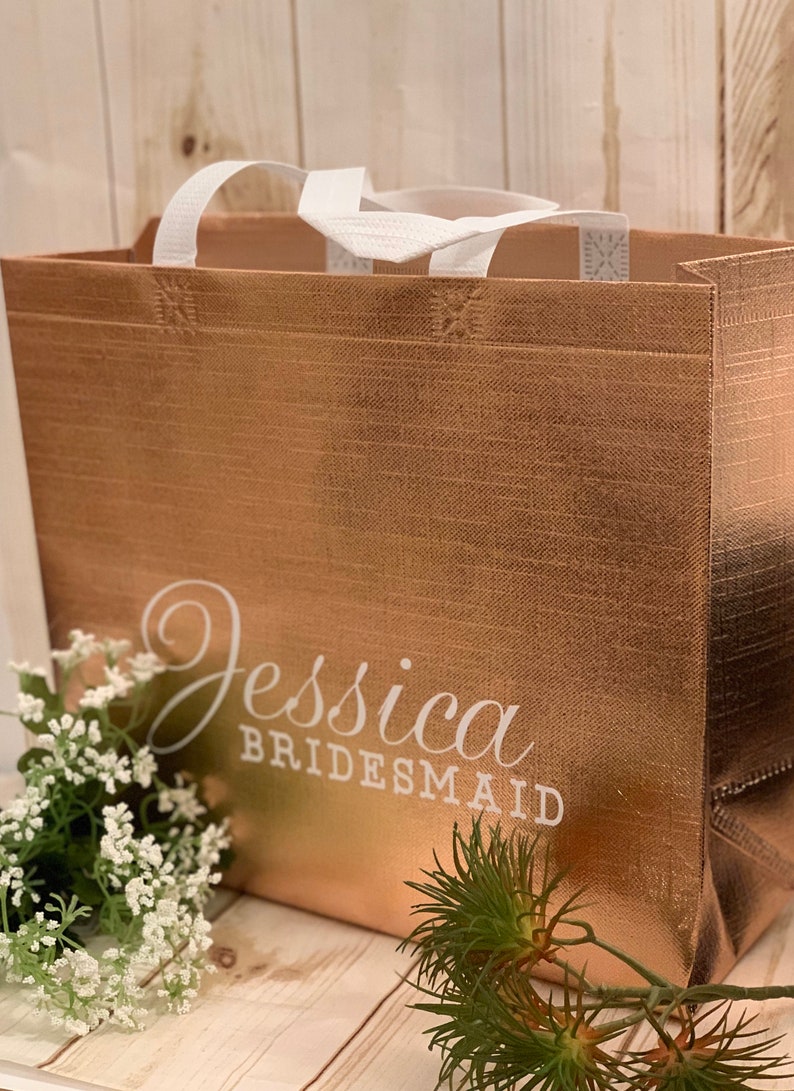 Puede incluir: Una bolsa de tela reutilizable de color oro rosa con asas blancas y el texto "Jessica Bridesmaid" impreso en la parte delantera.