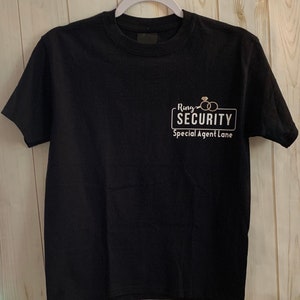 May include: Black t-shirt with the text "Ring Security Special Agent Lane" in white. The design includes two wedding rings. The shirt is hanging on a white hanger.