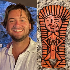 May include: A wooden carving of a person in a sarcophagus. The carving is painted in shades of brown and black. The person's face is smiling.