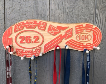 May include: A wooden shoe-shaped medal holder with a red outline and the numbers "26.2" and "10K" engraved on it. The holder has several white pegs for hanging medals.