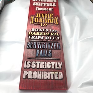May include: A red wooden sign with white, yellow, and blue lettering that reads "ATTENTION SKIPPERS The Use of JUNGLE NAVIGATION CO. BOATS for DAREDEVIL TRIPS OVER SCHWEITZER FALLS IS STRICTLY PROHIBITED".