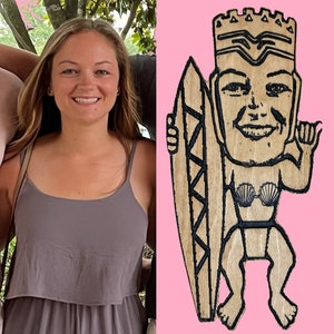 May include: A wooden cutout of a person holding a surfboard. The person is wearing a crown and a seashell bikini top. The cutout is painted in brown and black.