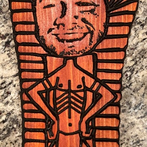 May include: A wooden cutout of a person in a pharaoh's headdress. The cutout is painted in shades of brown and black. The person is smiling and has a muscular physique.