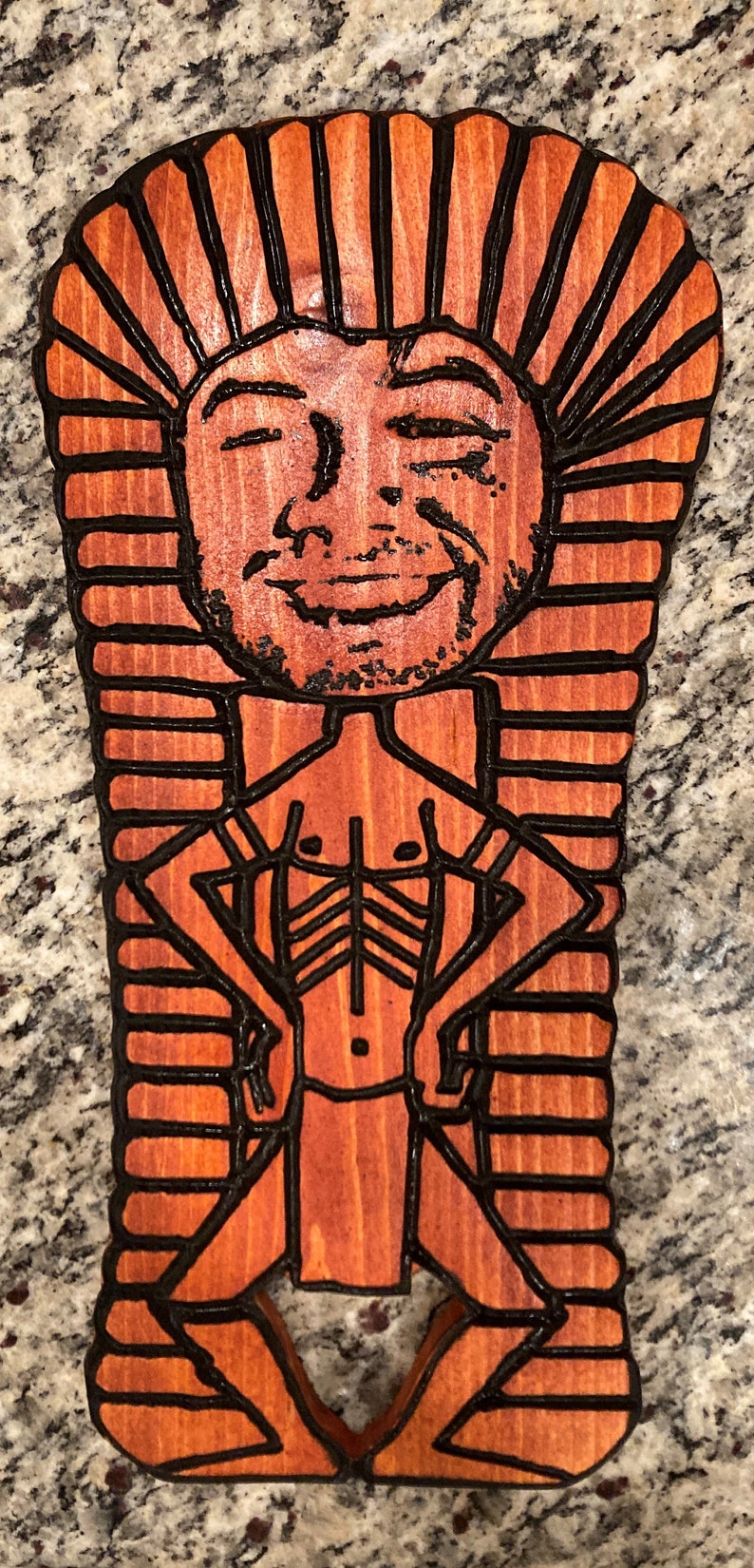 May include: A wooden cutout of a person in a pharaoh's headdress. The cutout is painted in shades of brown and black. The person is smiling and has a muscular physique.