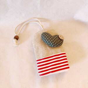 May include: A small, gray, heart-shaped fabric pouch with white polka dots and a red and white striped bottom. The pouch has a brown wooden bead and a loop of twine at the top.