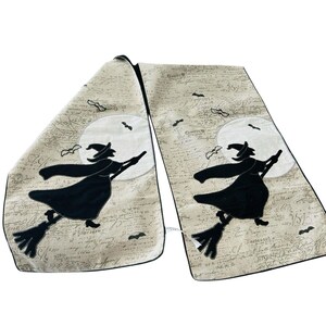 May include: Beige Halloween table runner featuring a black witch silhouette on a broomstick, flying in front of a full moon. The design includes bats and script-like text. The runner is made of a soft fabric.