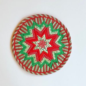 Op de afbeelding: Een rood en groen gestikt kerstster ornament met een witte achtergrond. De ster is gemaakt van stofstroken en heeft een rode rand.