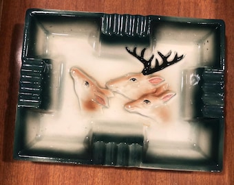 Op de afbeelding: Een rechthoekige keramische asbak met een reliëf van drie hertenkoppen in het midden. De asbak heeft een crèmekleurige basis met donkergroene gradiëntranden en vier geïntegreerde inkepingen voor het neerleggen van voorwerpen langs de rand.