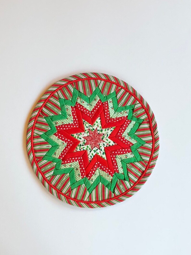 Op de afbeelding: Een rood en groen gestikt kerstster ornament met een witte achtergrond. De ster is gemaakt van stofstroken en heeft een rode rand.