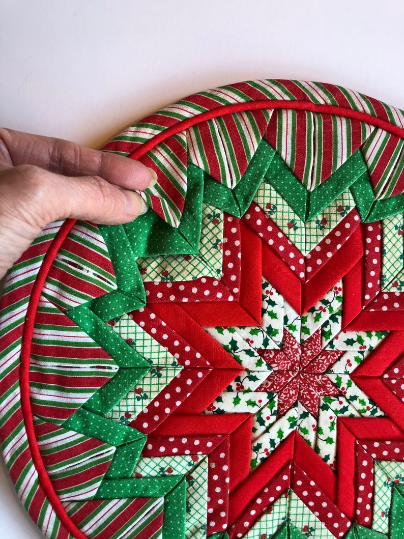 Op de afbeelding: Een rode en groene kerst-theedoek met een stervormig ontwerp. De theedoek is gemaakt van stofresten in verschillende patronen, waaronder stippen, strepen en hulst.