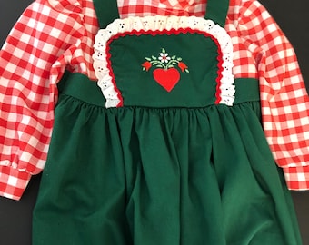 Op de afbeelding: Een groene en rode ruitjesjurk met een witte kantversiering en een rood hart geborduurd op de voorkant. De jurk heeft een witte kraag en rode ruitjesmouwen.