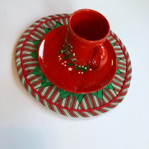 Op de afbeelding: Rode en groene kerst-thema stoffen placemat met een rode schotel en een rode mok met een wit kerstboomontwerp.