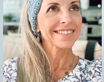 May include: A woman with long, light blonde hair is wearing a light blue crocheted headband. She is smiling and looking up. She is wearing a white and blue floral print blouse and a silver necklace with a small flower pendant.