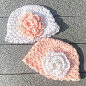 Puede incluir: Dos gorros de ganchillo con flores. Un gorro es blanco con una flor de melocotón, y el otro es melocotón con una flor blanca.