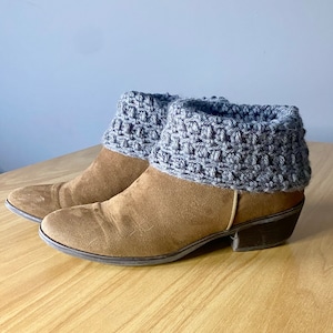 Puede incluir: Un par de botines de ante marrón con puños de ganchillo gris. Las botas tienen un tacón bajo y una puntera redonda.