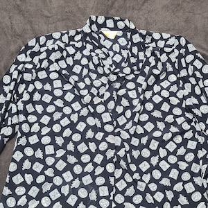 May include: A black long-sleeved blouse with a white pattern of repeating small images. The blouse has a button-up front and a bow tie at the neck.