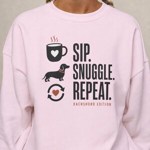 Könnte beinhalten: Ein hellrosa Sweatshirt mit Rundhalsausschnitt und einem Grafikdruck, der eine schwarze Kaffeetasse, einen schwarzen Dackel und ein Recyclingsymbol mit Herz zeigt. Der Text lautet SIP. SNUGGLE. REPEAT. DACHSHUND EDITION in Schwarz und Orange.