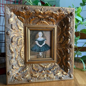 Puede incluir: Un pequeño retrato pintado de un niño con el pelo largo y un vestido verde oscuro, colocado dentro de un marco ancho, ornamentado y de color dorado con patrones de hojas y flores en relieve. La obra es rectangular y muestra una figura sentada.