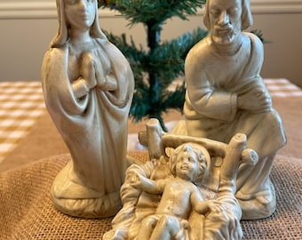 Puede incluir: Un belén de cerámica vintage que representa a María, José y el niño Jesús. Las figuras están pintadas en color crema y tienen un aspecto rústico y envejecido.