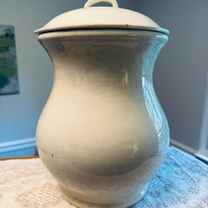 May include: A cream-colored ceramic jar with a rounded body and a matching lid featuring a small arched handle. The surface has a smooth, glossy finish with subtle variations in tone and a visible horizontal line near the base of the jar.