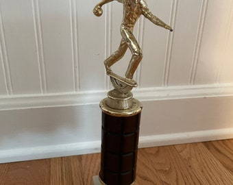 May include: A gold-colored bowling trophy featuring a figure of a person bowling. The trophy has a brown base with a marble-like top.