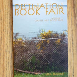 May include: A book fair poster with the text "DESTINATION BOOK FAIR" in gold letters. The poster features a landscape with a chain-link fence, yellow flowers, and a body of water. The text "SEATTLE ART BOOK FAIR" is also visible.
