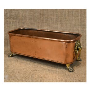 May include: A rectangular copper planter with a polished finish. It features brass lion's head handles and brass claw feet. The interior shows signs of age. The planter is set against a neutral background.