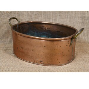 May include: Oval copper planter with brass handles. The planter has a polished exterior and a slightly aged patina, showing signs of use. The interior has a dark, aged appearance. The handles are attached to the sides.