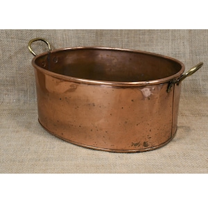 May include: Oval copper planter with brass handles. The planter has a polished finish and is empty. The handles are curved and attached to the sides. The planter is on a burlap surface.
