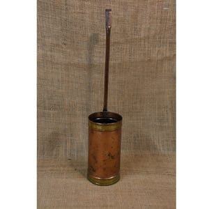 May include: A vintage copper and brass container with a long handle. The cylindrical body has a tarnished copper finish with brass accents at the top and bottom. The handle extends upwards, ending in a flat, rectangular shape.