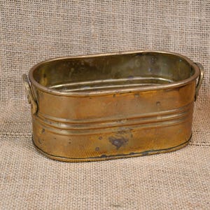 May include: An antique brass oval planter with two small handles. The planter shows signs of age and wear. The shape is rectangular with rounded ends. The background is a neutral colour, and the planter is made of brass.