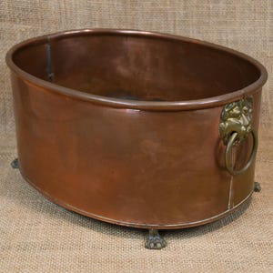 May include: Oval copper planter with a polished finish. The planter features a lion's head ring handle and rests on three claw feet. The copper has a warm, reddish-brown hue, and the planter appears to be antique.