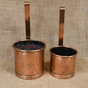 May include: Two antique copper milk measuring cups with long handles. The cups are cylindrical with a slightly flared rim and the word "Milk" is stamped on the side. The copper has a warm, aged patina.