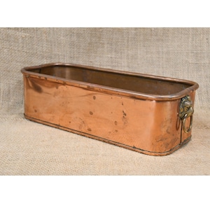 May include: A rectangular copper planter with a weathered patina. The planter has a rolled rim and a decorative lion's head handle on one end. The copper has a warm, reddish-brown hue, and the planter appears to be vintage.