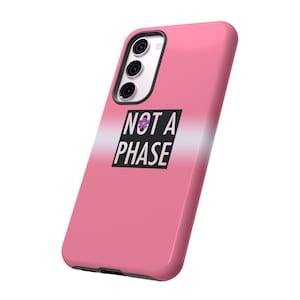 Puede incluir: Funda de teléfono rosa con un degradado blanco y rosa claro y el texto "NOT A PHASE" en negro con una flor morada.