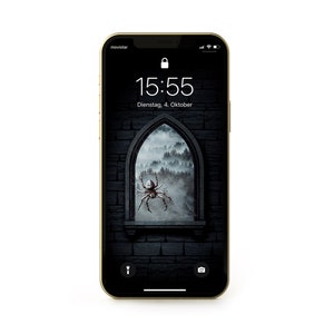 Puede incluir: Un iPhone dorado con una pantalla negra que muestra un fondo de pantalla de estilo gótico oscuro. El fondo de pantalla presenta una araña en una ventana con un bosque brumoso en el fondo. La hora es 15:55 y la fecha es Dienstag, 4. Oktober.