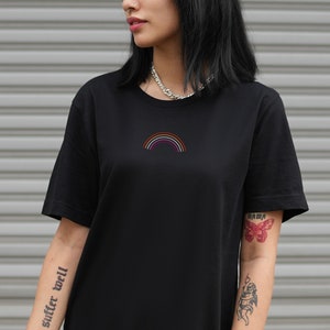 Puede incluir: Una camiseta negra con un pequeño gráfico de arcoíris en la parte delantera. El arcoíris está formado por seis colores: rojo, naranja, amarillo, verde, azul y morado. La camiseta tiene cuello redondo y mangas cortas.