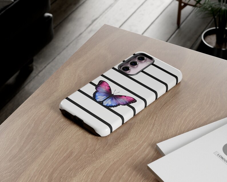 K&ouml;nnte beinhalten: Eine wei&szlig;e Handyh&uuml;lle mit schwarzen Streifen und einem bunten Schmetterlingsdesign. Die H&uuml;lle hat einen Ausschnitt f&uuml;r das Kameraobjektiv und einen erh&ouml;hten Rand an den Kanten.