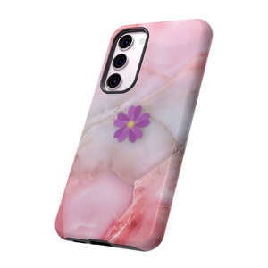 Puede incluir: Una funda de teléfono de mármol rosa con un diseño de flor morada. La funda tiene una protuberancia de cámara elevada y un borde negro.