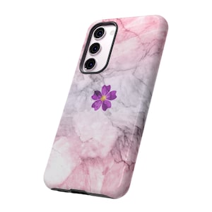 Puede incluir: Una funda de teléfono de mármol rosa y gris con un diseño de flor morada. La funda tiene una protuberancia de cámara elevada y un borde negro.