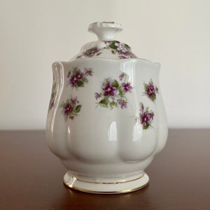Peut inclure: Sucrier en porcelaine blanche avec un couvercle, décoré d'un motif floral de violettes violettes et blanches. Le bol a une bordure dorée autour de la base et du couvercle.