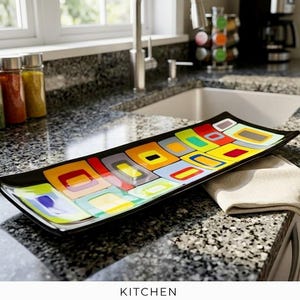 Puede incluir: Una bandeja de servicio rectangular de vidrio con un patrón geométrico multicolor de cuadrados anidados en tonos rojo, amarillo, azul, verde y gris, sobre un borde negro. La bandeja tiene un acabado brillante y bordes curvos.
