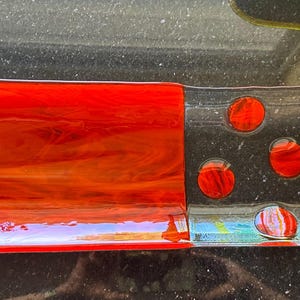 May include: A rectangular fused glass dish with a striking design. It features a large section of fiery red glass, a section with red circles on a black background, and another red section. The glass has a glossy finish, with a modern aesthetic.