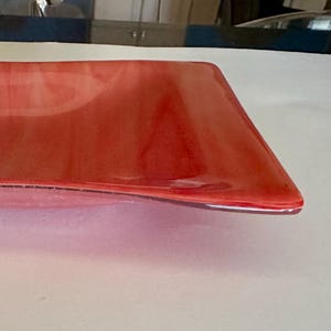 Może przedstawiać: Prostokątny, czerwony szklany talerz do serwowania o lekko zakrzywionym kształcie. Talerz ma błyszczące wykończenie i wydaje się być ręcznie robiony. Kolor jest żywy, półprzezroczysty czerwony, odpowiedni do serwowania przystawek lub małych potraw.