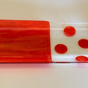 Puede incluir: Una fuente rectangular de vidrio con forma curva. La fuente presenta secciones de vidrio rojo vibrante y transparente. Lunares rojos están dispuestos en la sección transparente. La fuente mide aproximadamente 30 cm de largo.