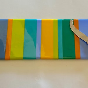 May include: A rectangular serving board with vertical stripes in orange, blue, yellow, and green. A silver cheese knife with a dotted handle rests on the board. The board is made of glass or a similar material.