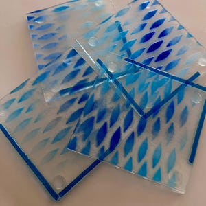 May include: A set of four square glass coasters featuring a repeating pattern of blue, leaf-shaped marks on a translucent, frosted background. Each coaster has rounded corners and a distinct blue border along the edges, creating a cohesive set.
