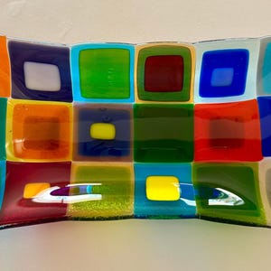 May include: A rectangular fused glass tray featuring a vibrant grid pattern of multicolored squares. The design includes blocks of red, orange, yellow, green, blue, and white, arranged in a repeating mosaic style with smooth, glossy surfaces.